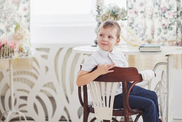 Niño pequeño en pantalones vaqueros azules está sentado en la silla de madera en la sala. Hace la tarea. De vuelta a la escuela