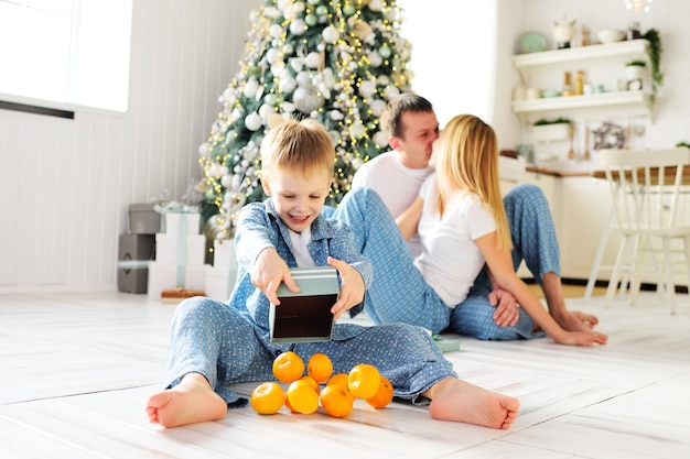 Un niño pequeño en pijama azul sentado en un piso cálido abre una caja de regalo con padres mandarinas y un árbol de Navidad. Celebración de Navidad.