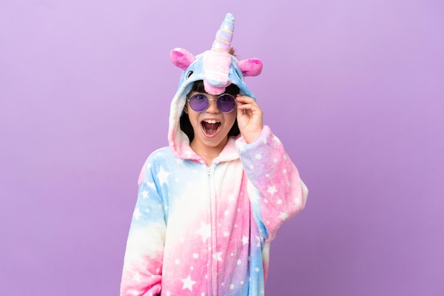 Niño pequeño con un pijama de unicornio aislado de fondo morado con gafas y sorprendido