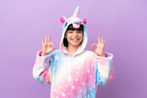 Niño pequeño con un pijama de unicornio aislado en un fondo morado que muestra un signo de ok con los dedos