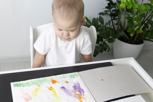 Un niño pequeño pinta con pinturas de acuarela en una mesa junto a una flor verde