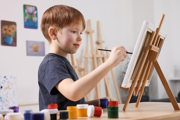Niño pequeño pintando en la mesa en el estudio usando un caballete para sostener el lienzo