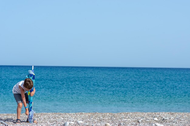 Niño pequeño plegable sombrilla en una hermosa playa de guijarros con océano azul detrás de él