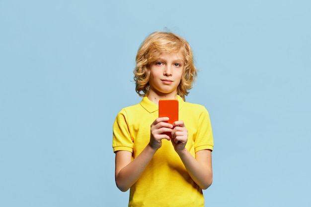 Niño pequeño posando en camiseta amarilla tomando fotos con teléfono móvil contra estudio azul