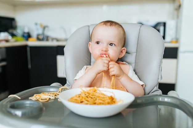 El niño pequeño que come espaguetis parece hambriento y la salsa de pasta se ensucia alrededor de su cara mientras toma ...