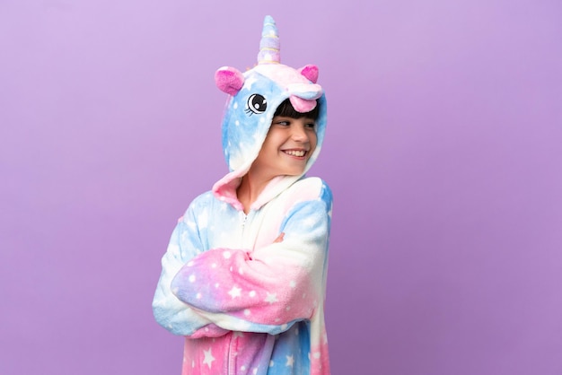 Niño pequeño que lleva un pijama de unicornio aislado en un fondo morado con los brazos cruzados y feliz