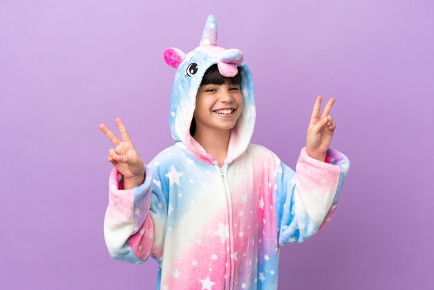 Niño pequeño que lleva un pijama de unicornio aislado en un fondo morado que muestra el signo de la victoria con ambas manos