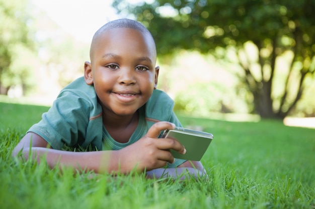 Niño pequeño que miente en la hierba que sostiene la cámara digital que sonríe en la cámara