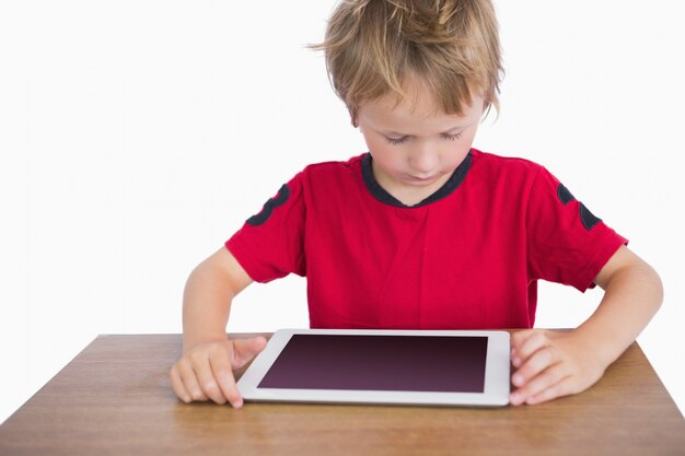 Niño pequeño que se sienta en el escritorio y que mira la tableta digital