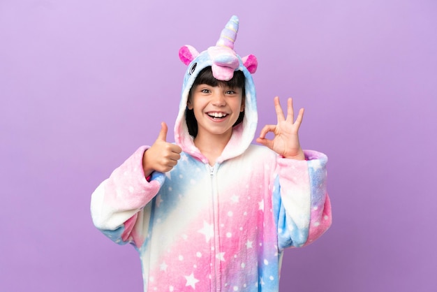 Niño pequeño que usa un pijama de unicornio aislado en un fondo morado que muestra el signo de ok y el gesto del pulgar hacia arriba