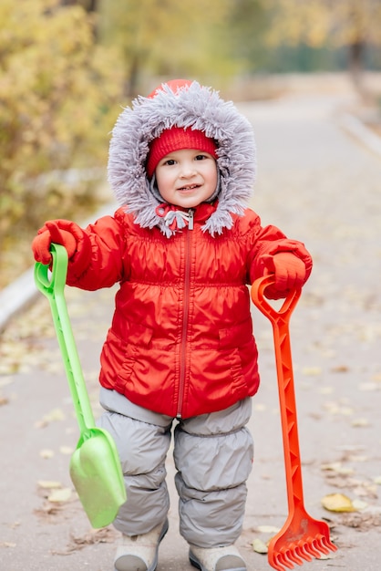 Un niño pequeño con un rastrillo y una pala camina por el parque