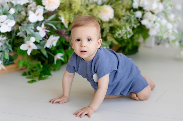 Un niño pequeño recién nacido con un traje azul se sienta en el suelo del dormitorio rodeado de hermosas flores. Mira a la cámara. Maqueta de productos para bebés.