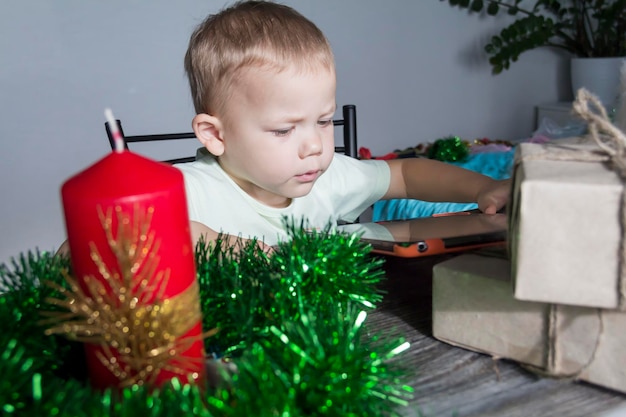 Un niño pequeño con regalos de Navidad está sentado en una mesa de madera de Año Nuevo con una tableta en las manos