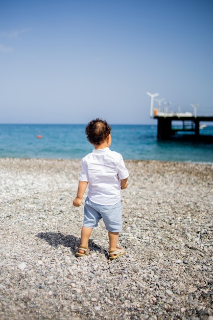 Niño pequeño rizado juega en la playa de guijarros cerca del mar azul