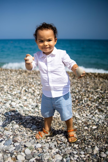 Niño pequeño rizado juega en la playa de guijarros cerca del mar azul