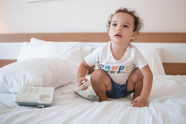 Niño pequeño rizado jugando con un teléfono con cable en la cama