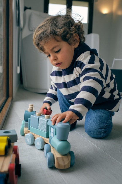Niño pequeño con ropa casual sentado en el suelo y jugando con el tren de juguete mientras pasa tiempo en casa