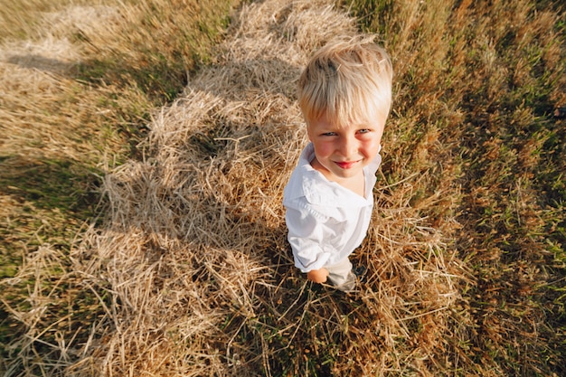 Niño pequeño rubio que juega el heno en el campo. verano, clima soleado, agricultura.