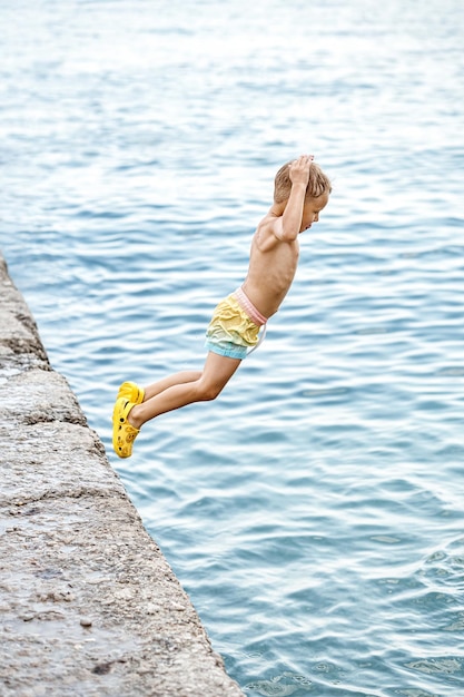 Foto niño pequeño salta volando desde el muelle en una postura divertida al agua de mar contra el paisaje marino borroso
