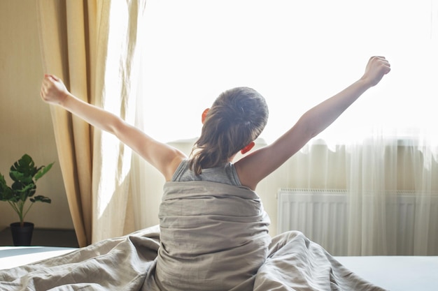 Niño pequeño sentado y estirándose en la cama en casa por la mañana en un fondo de ventana