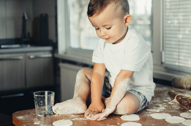 Un niño pequeño sentado en una mesa cubierta de harina con masa en sus manos sonriendo