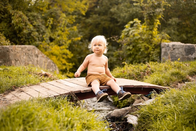 Niño pequeño sentado en pantalones cortos en el puente sobre el arroyo en otoño o parque de verano con árboles dorados