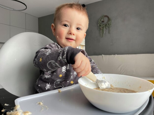 Un niño pequeño sentado en una silla alta come avena por sí mismo con una cuchara