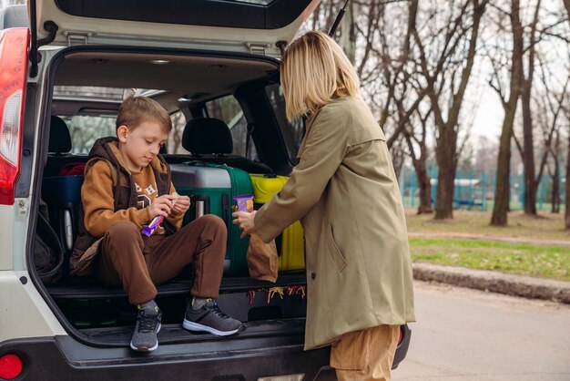 Niño pequeño sentado con su madre en el baúl del auto lleno de equipaje comiendo dulces de chocolate