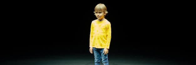 Un niño pequeño, serio, sincero, caucásico, de seis años, con el pelo largo y rubio, está parado alto en el estudio iluminado por una pancarta de luz blanca y amarilla brillante