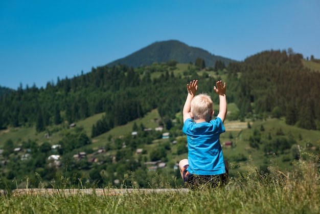 El niño pequeño se sienta en las montañas con las manos levantadas. Vista trasera