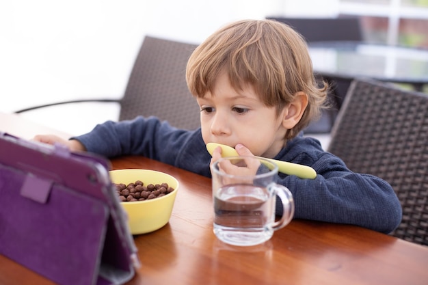 Un niño pequeño se sienta en una silla en la mesa y mira dibujos animados en una tableta lamiendo una cuchara Un niño en edad de jardín de infantes come bolas de chocolate con leche y bebe agua de una taza Adicción a los aparatos para el desayuno listo