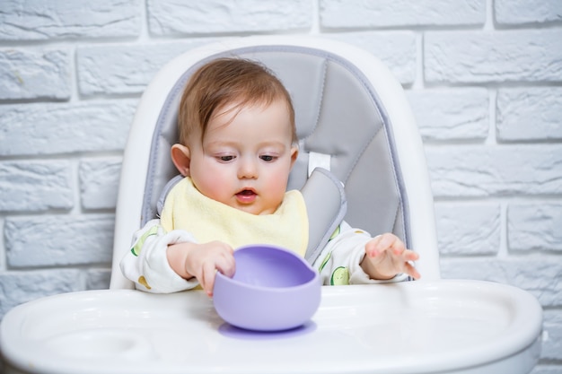 Un niño pequeño se sienta en una trona y come la comida de un plato con una cuchara. Utensilios de silicona para bebés para alimentar a los bebés