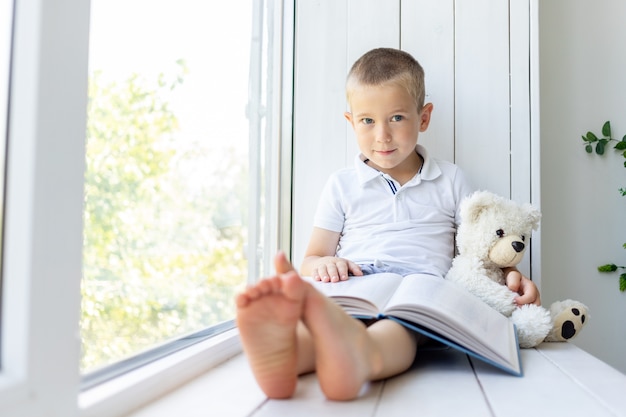 Un niño pequeño se sienta en la ventana con un libro y un oso blando.