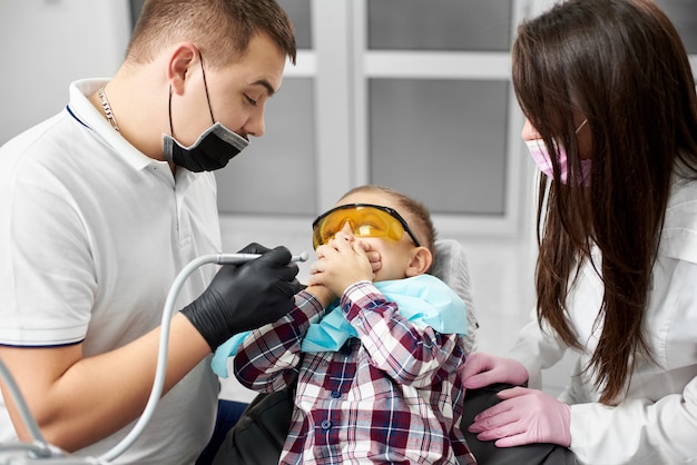 Un niño pequeño en un sillón dental cierra la boca con las manos, temiendo a un dentista que tiene un taladro en la mano.