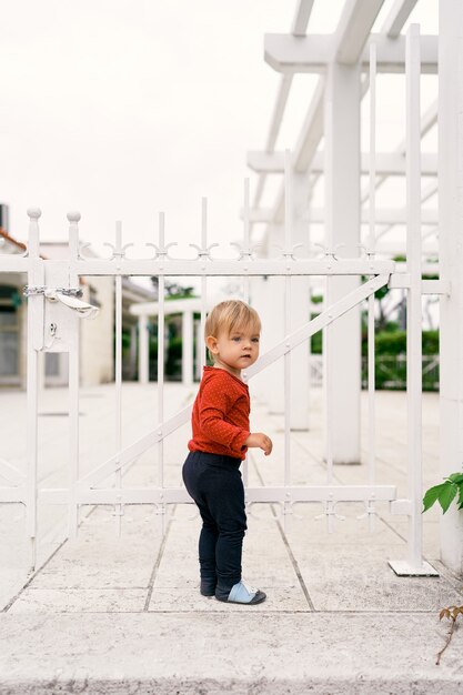 Niño pequeño se para sobre las losas y sostiene su mano a una puerta de metal blanco cerrada