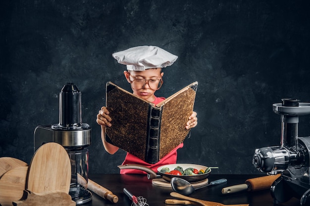 Un niño pequeño con sombrero y gafas de chef está leyendo un libro de recetas y listo para cocinar verduras.