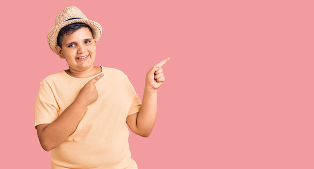 Niño pequeño con sombrero de verano y traje de baño hawaiano sonriendo y mirando a la cámara apuntando con dos manos y dedos a un lado