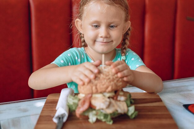 Un niño pequeño y sonriente con el pelo rubio está buscando con apetito una jugosa hamburguesa enorme en un café. Retrato. Niños.