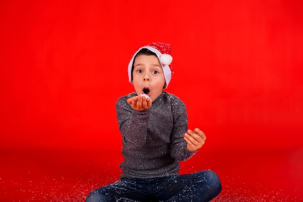 El niño pequeño sopla nieve artificial de su mano sobre un fondo rojo.