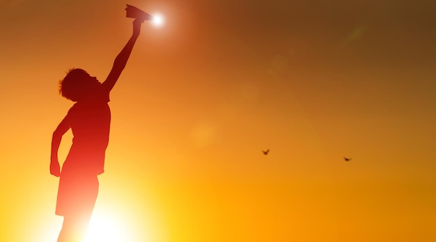 Un niño pequeño sostiene un avión de papel en la mano al atardecer Un niño levantó la mano hacia el cielo y juega con origami por la noche en el lago Silhouette