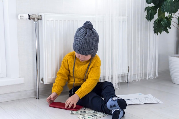 Un niño pequeño con un suéter amarillo y un sombrero está contando dinero y estudiando las facturas de calefacción cerca de un calentador con termostato