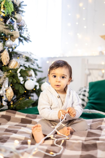 Un niño pequeño con un suéter de punto está sentado en la cama contra el fondo de un árbol de Navidad decorado festivo en casa, el niño está celebrando Navidad y Año Nuevo en casa