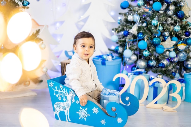 Un niño pequeño con un suéter de punto está sentado en un trineo contra el fondo de un árbol de Navidad decorado festivo en casa sonriendo Año Nuevo 2023 Navidad