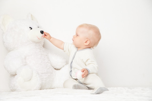 Niño pequeño en traje blanco tocando la nariz del oso de peluche sobre fondo blanco.