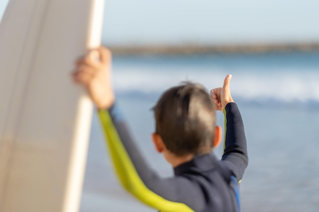 Un niño pequeño con traje de neopreno muestra los pulgares hacia arriba mirando la vista trasera de la tabla de surf