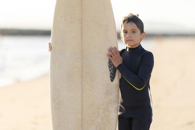 Un niño pequeño con traje de neopreno sosteniendo una tabla de surf en la orilla del mar