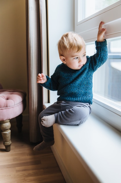 Niño pequeño travieso con cabello rubio y ojos azules, se sienta en el alféizar de la ventana, mira por la ventana