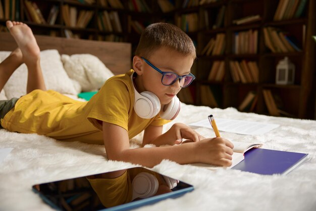 Foto niño pequeño usando un gadget digital moderno estudiando en casa mientras está acostado en la cama educación cómoda y infancia feliz