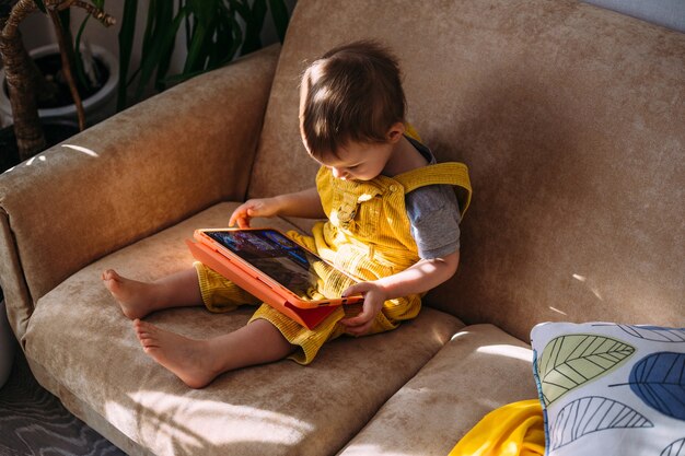 Un niño pequeño ve dibujos animados en una tableta mientras está sentado en el sofá solo en casa.