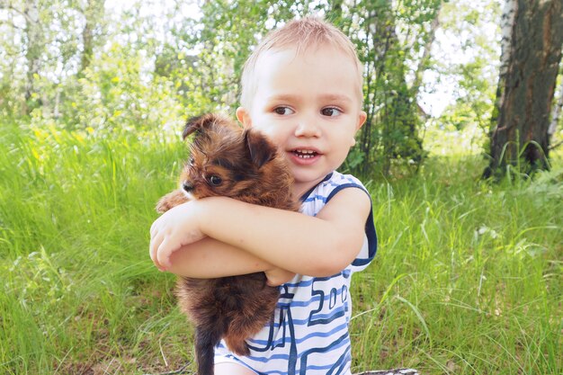 Un niño pequeño en el verano en el bosque se sienta en un tocón y abraza a un pequeño cachorro marrón.